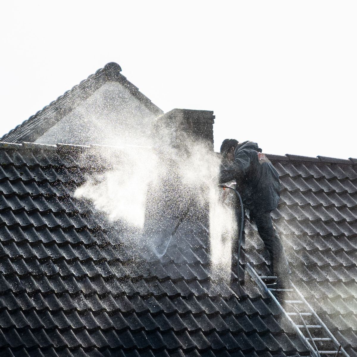 Technician on a ladder washing a dark tile roof